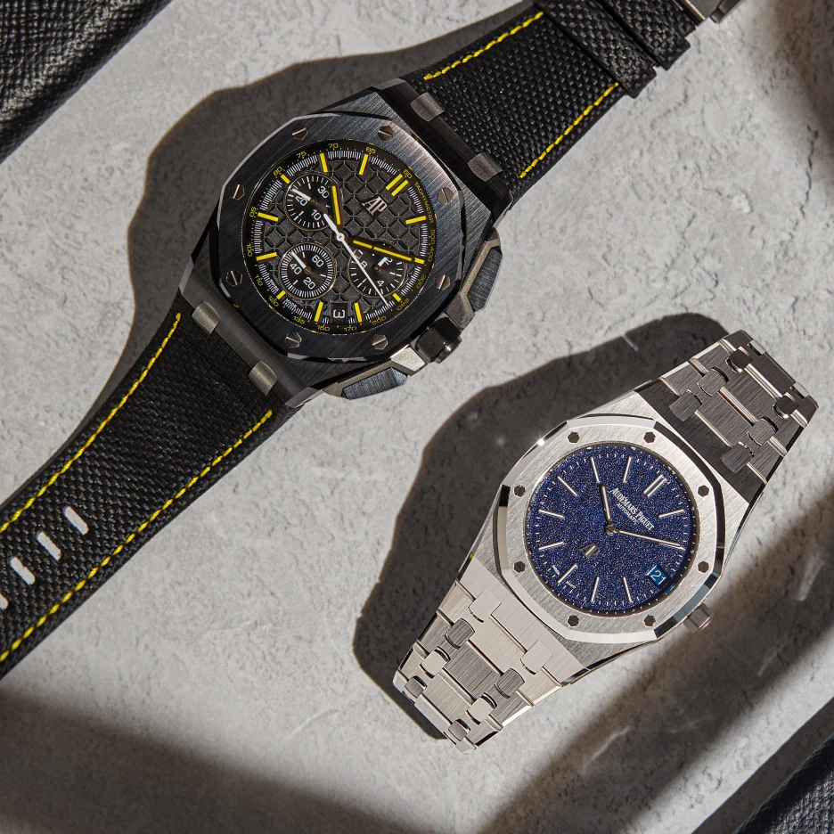 Two luxury wristwatches displayed on a textured surface, featuring metal and fabric straps with detailed watch faces.