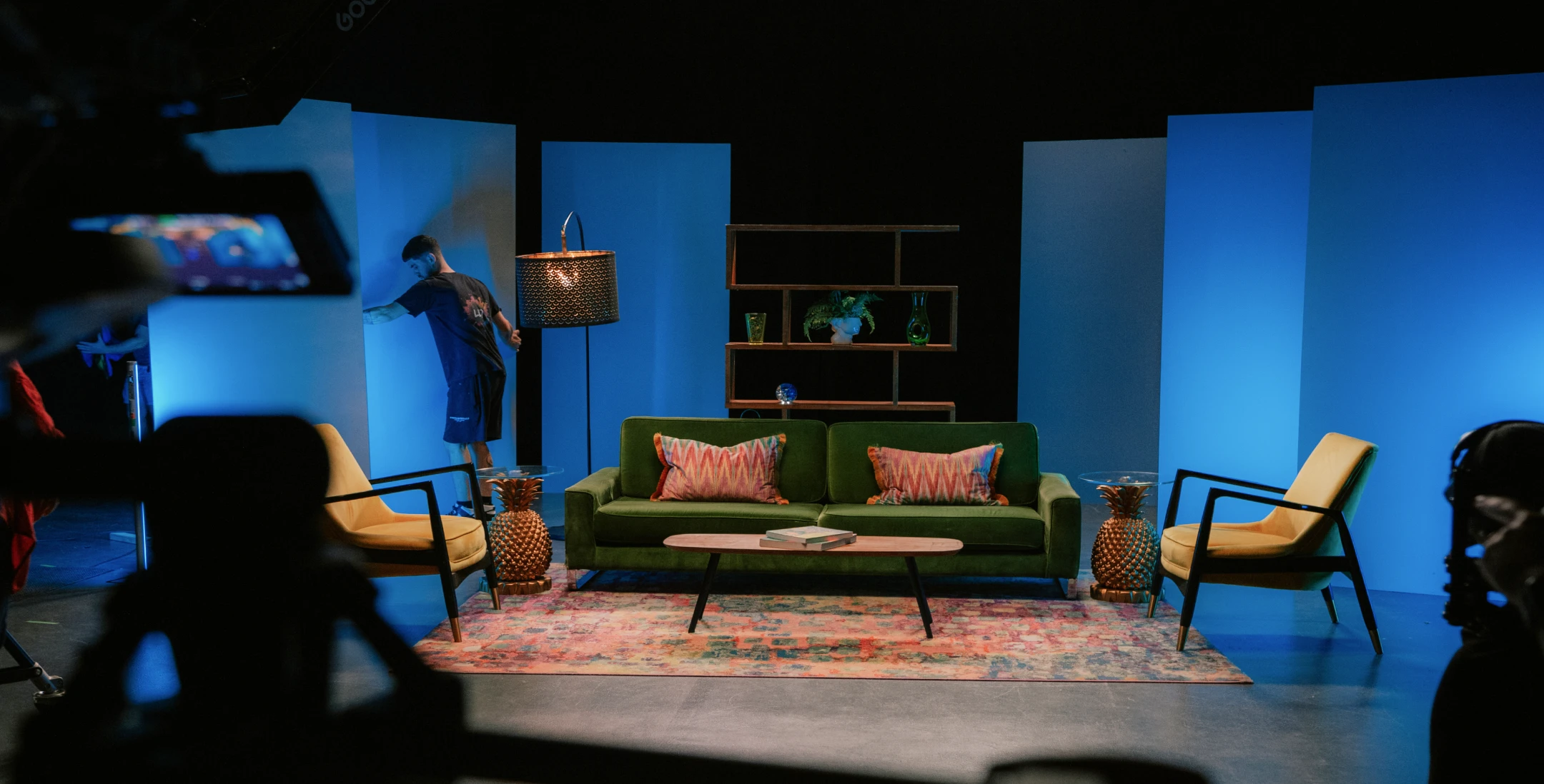 Behind-the-scenes studio set design featuring a styled living room stage with sofa, chairs, lighting and production cameras