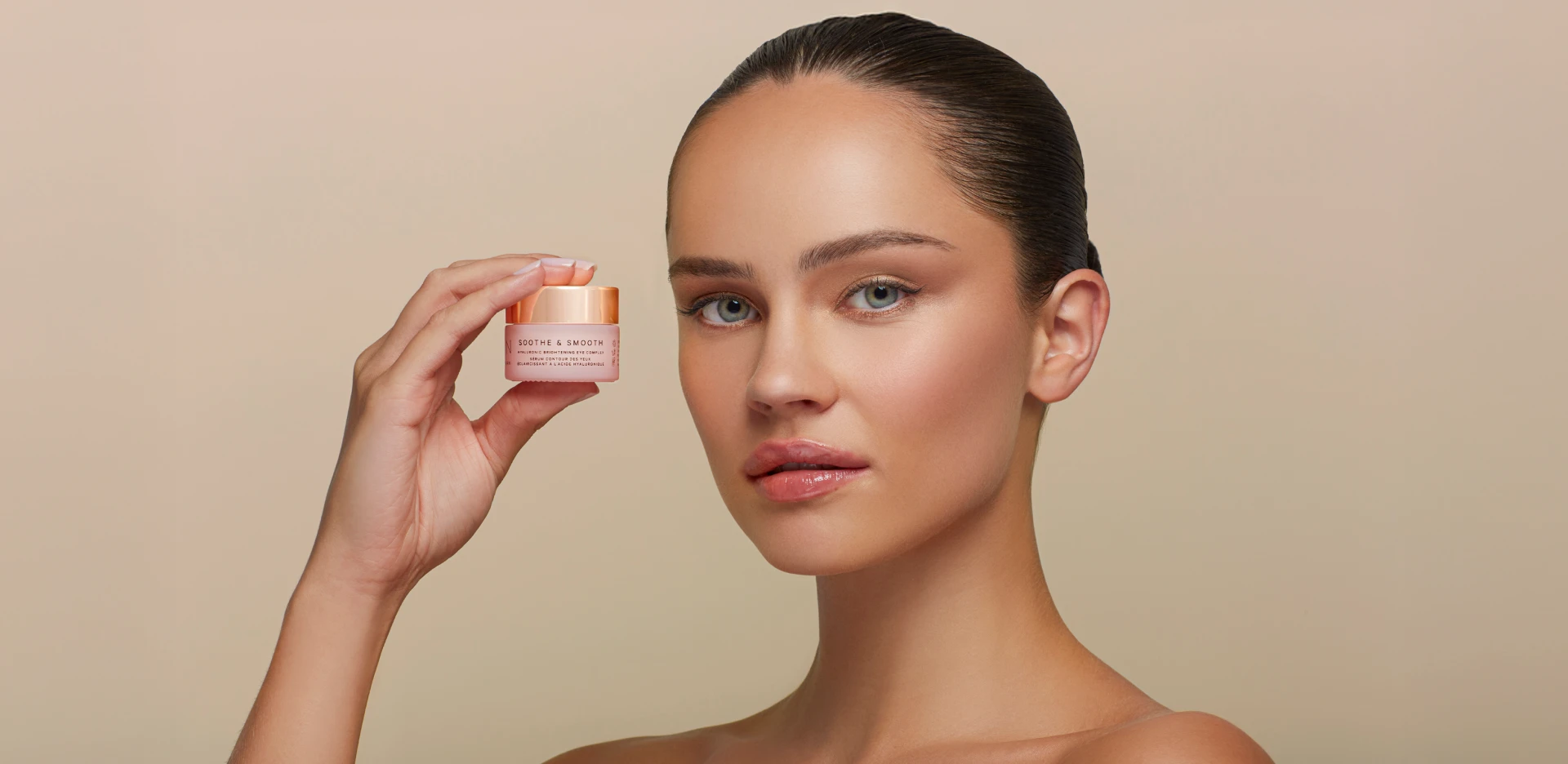 Woman holding a small jar of skincare cream next to her face against a neutral background, showcasing a cosmetic product.