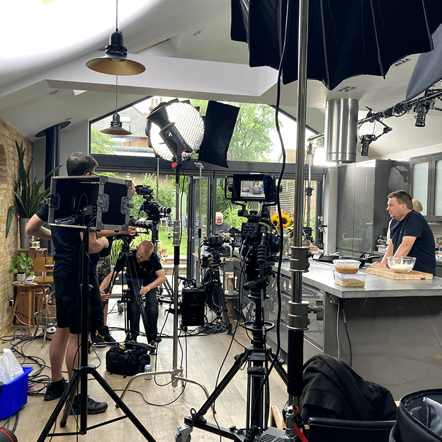 Video production crew filming a cooking scene in a modern kitchen with cameras and studio lighting equipment