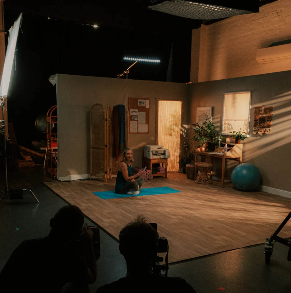 Studio set design for wellness video production with yoga instructor on mat, lighting rigs and cameras visible behind the scenes